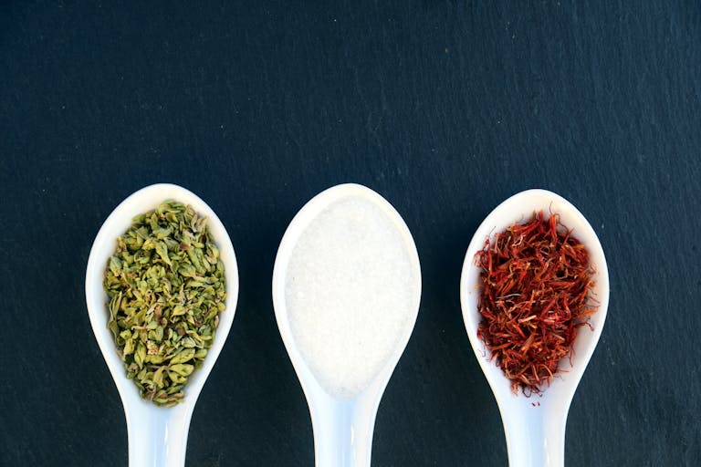 Three ceramic spoons with herbs, salt, and saffron on dark slate background.